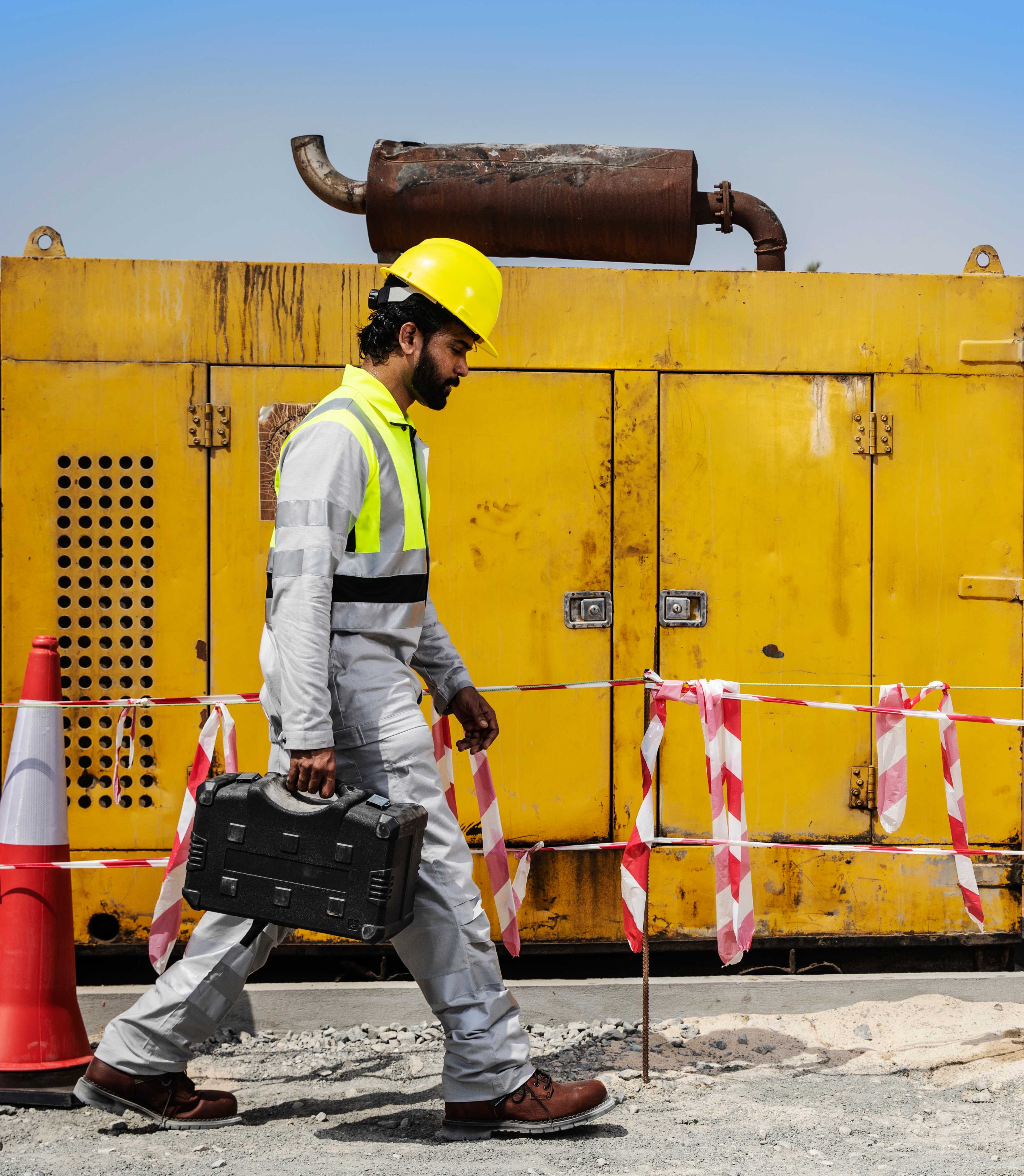 Construction worker wearing Shamal lightweight high-visibility workwear and hard hat walks past heavy equipment on a job site, showing reflective safety gear for extreme heat. Construction worker in Shamal high-visibility workwear walks on site carrying a tool case, wearing a safety helmet.