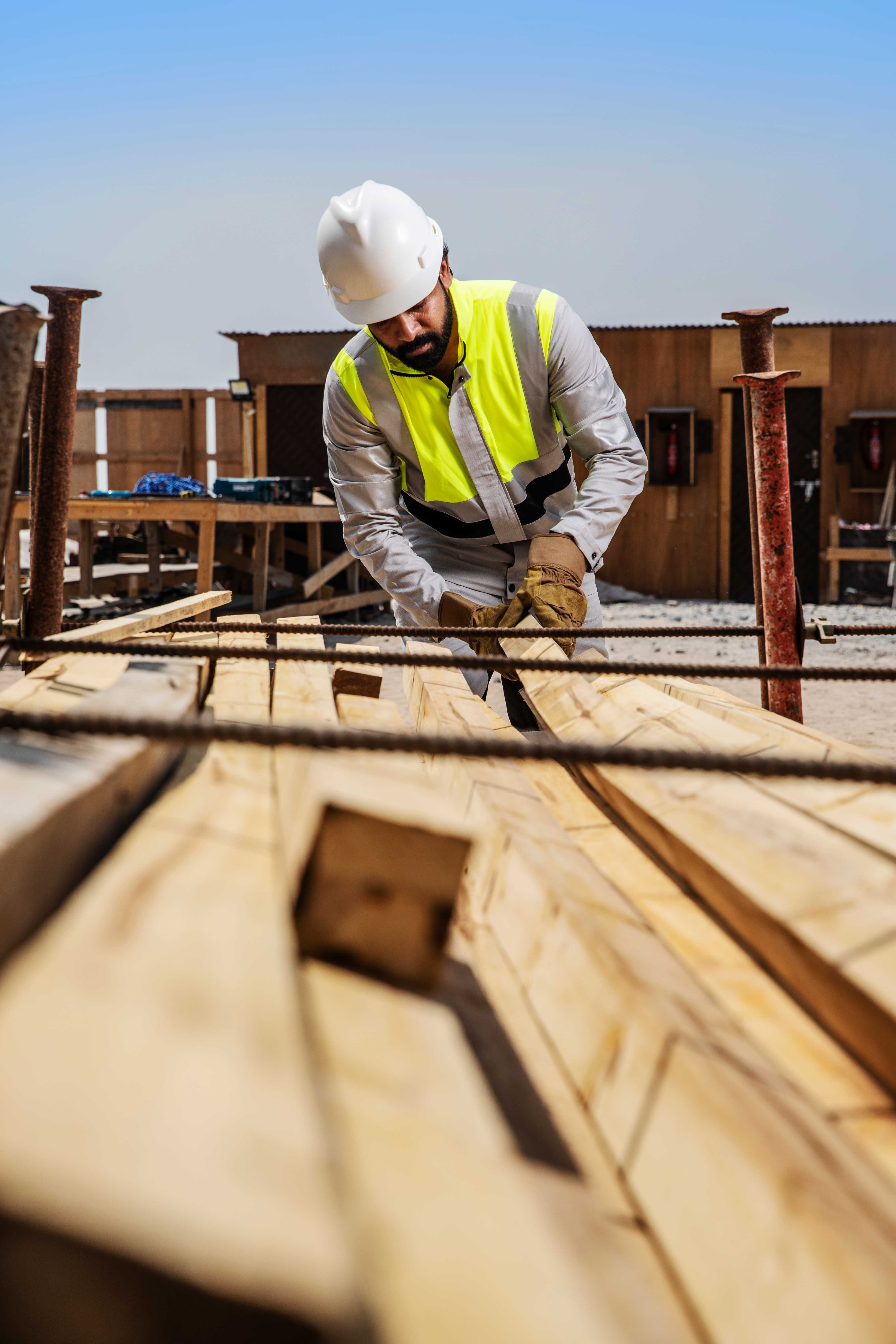 Construction worker wearing Shamal lightweight high-visibility workwear works with timber on a Middle East job site, showing reflective safety design for extreme heat conditions. Construction worker in Shamal high-visibility workwear and hard hat handles timber on a job site in bright daylight.