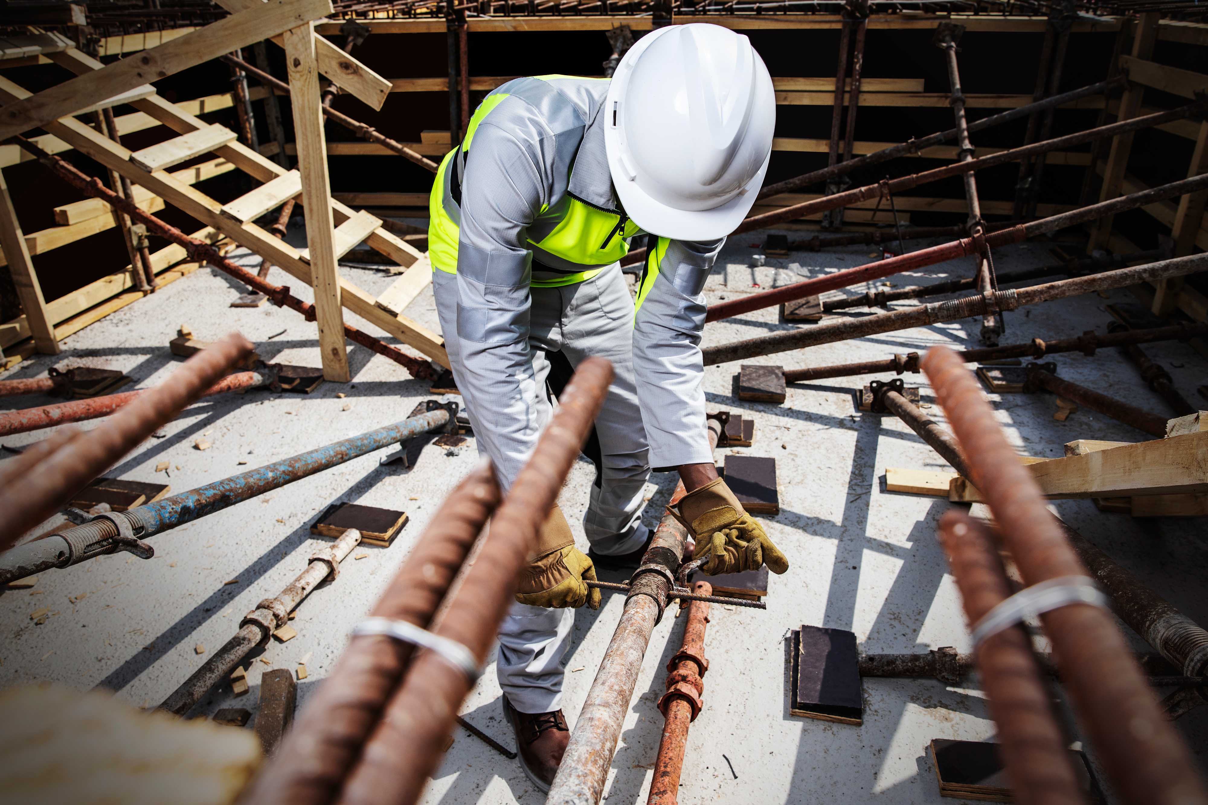 Construction worker wearing Shamal lightweight high-visibility workwear works with rebar and scaffolding on site, highlighting reflective safety gear designed for extreme heat. Construction worker in Shamal high-visibility workwear and hard hat adjusts rebar and scaffolding on a job site.