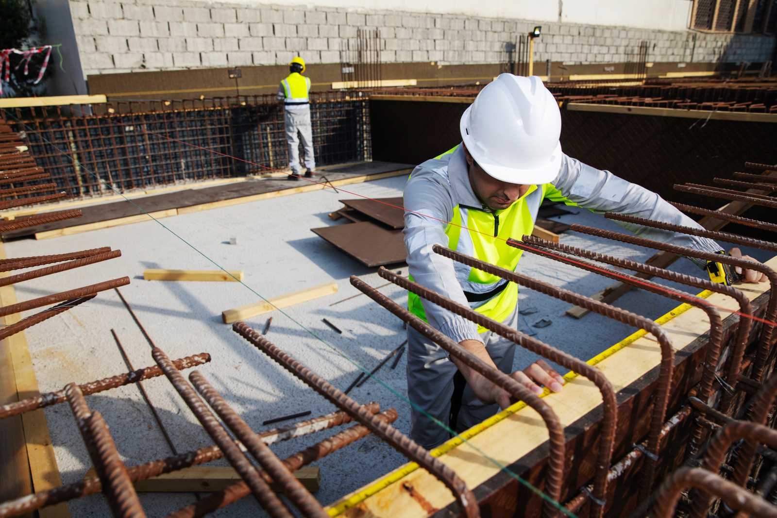 Construction worker wearing Shamal lightweight high-visibility workwear measures rebar and formwork on site, showing reflective safety gear designed for extreme heat conditions. Construction worker in Shamal high-visibility workwear measures rebar and formwork on a job site, with another worker in the background.