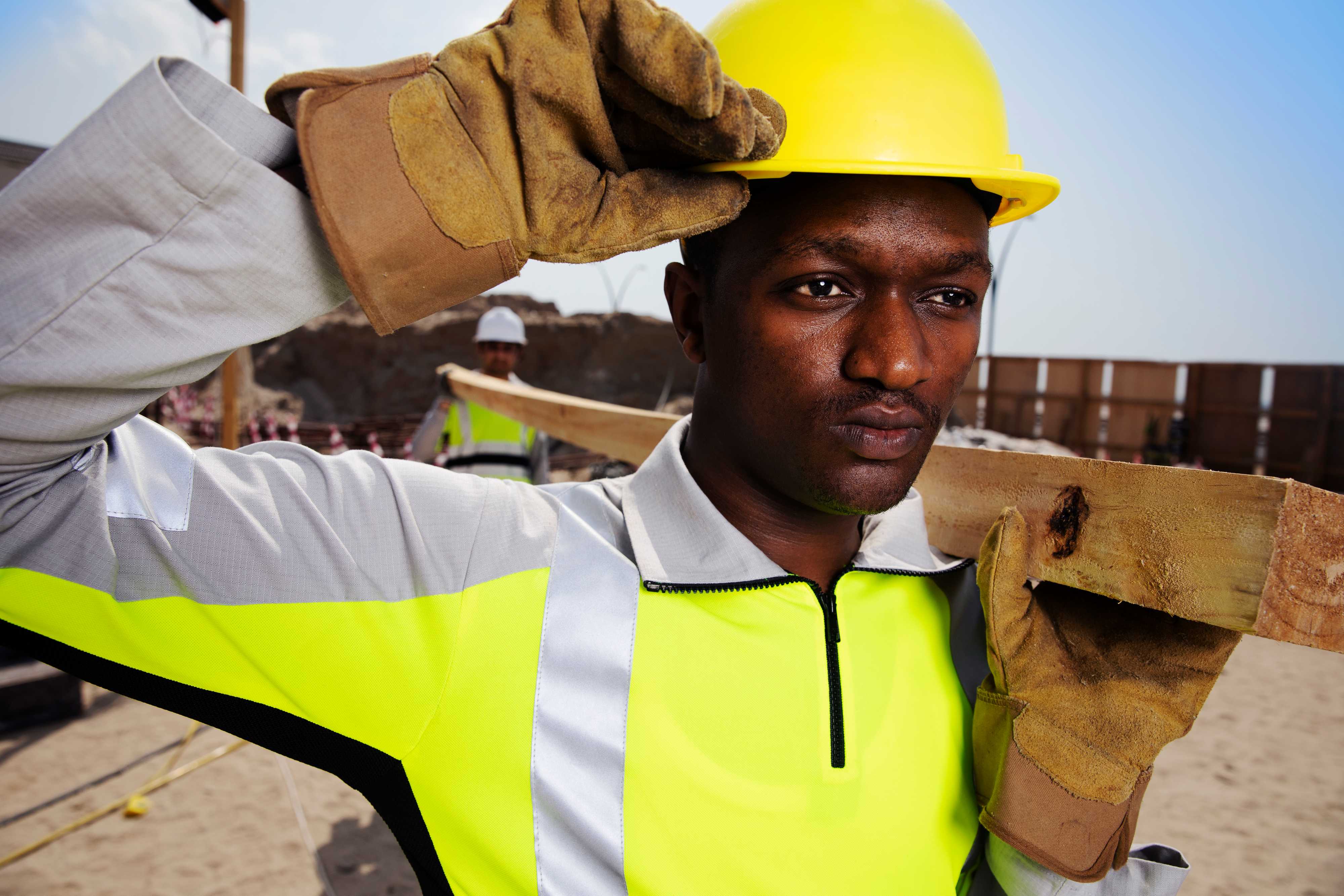 Shamal Workwear — lightweight construction workwear engineered for extreme heat. Construction worker wearing Shamal high-visibility workwear and hard hat on a site, carrying timber on his shoulder.