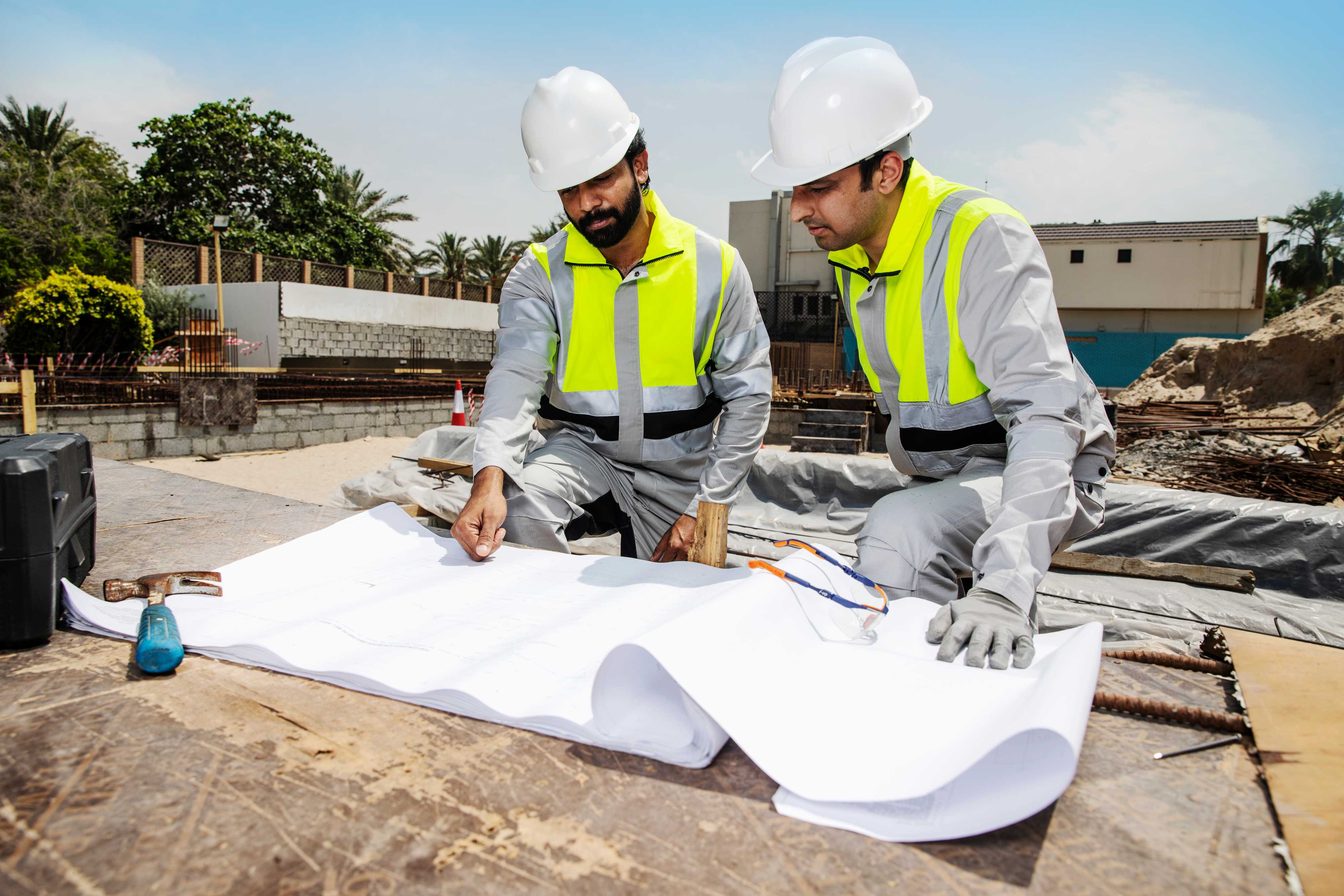 Construction workers wearing Shamal lightweight high-visibility workwear and hard hats review project plans on site, highlighting reflective safety gear for extreme climates. Two construction workers in Shamal high-visibility workwear review large site drawings on a worktable.